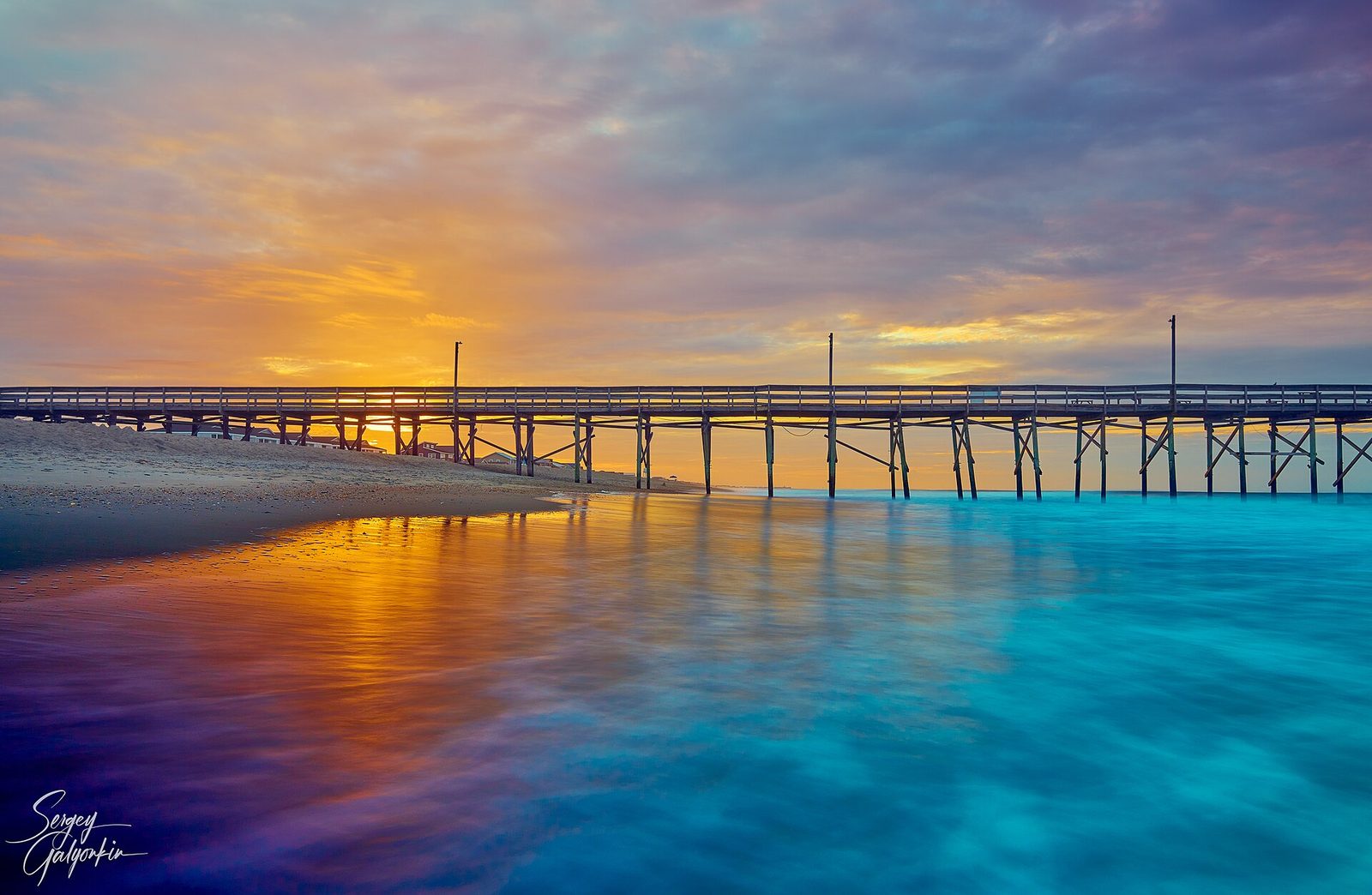 The Holden Beach fishing pier at dawn, with pink and gold sky reflected in the wet sand. The pier is now closed.