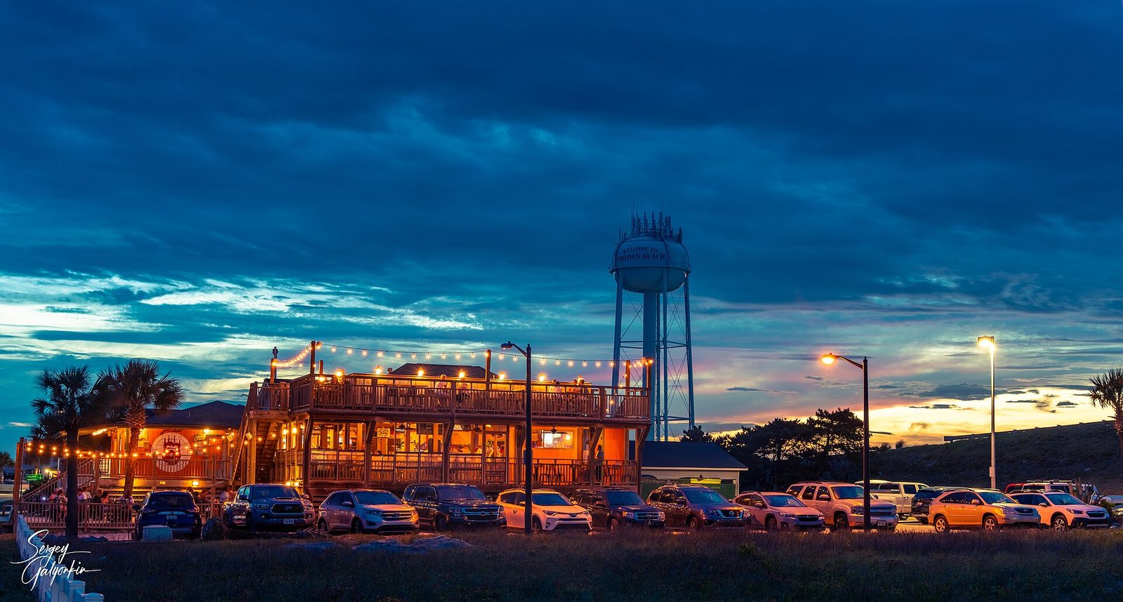 A lit-up restaurant and the Holden Beach water tower at dusk, with cars in the lot